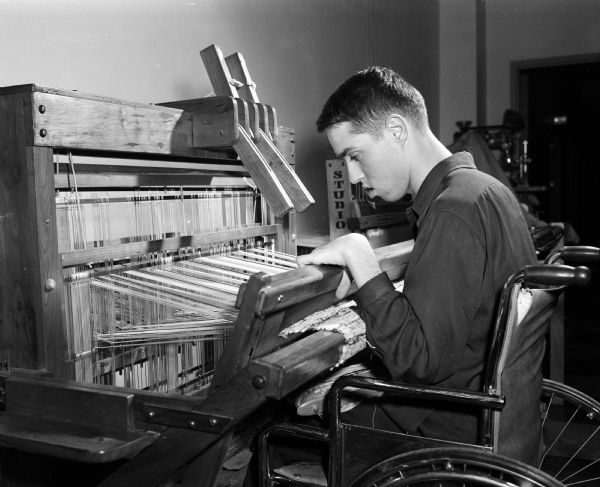 Dean Fiedler sits in a wheel chair while working at a loom. Fiedler wove rag rugs at the Homecrafters' Training Center at the Vocational School. The rugs were sold at the Homecrafters Shop at Barron's Department Store. Fiedler, a 'spastic' since birth, was then able to earn his own money.