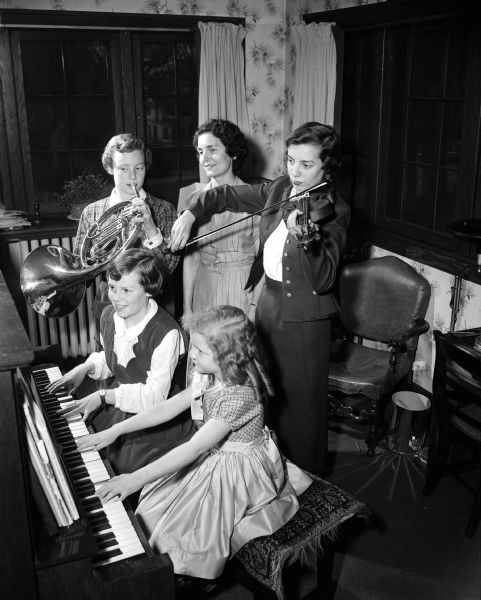 Group portrait of the Crownhart family gathered at a piano with french horn and violin. Clockwise from the bottom are: Gretchen, 6, seated at piano, Ginger, 11, also seated at piano, Sally, 15, playing a french horn, the mother, Marian looking on, and the oldest daughter, Maryann (Mrs. Warren Turner Jr.) playing a violin.