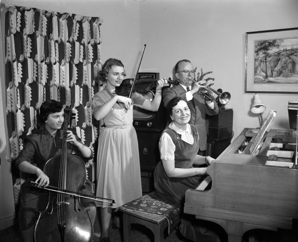 Portrait of the Fromm family playing music. From left, they are: Kathleen, 14, playing cello, Barbara, 18, playing violin, the mother Emily playing the piano while looking into the camera, and the father, Dr. Arno Fromm, playing his trumpet. The mother, Emily Fromm, had a weekly television program over WKOW-TV. In the background is a television set and a mid-century patterned curtain.