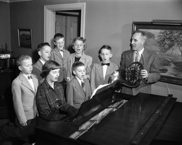 Group portrait of eight members of the Arthur Hasler family posed around a piano, singing. Seated at the piano are Sylvia, the oldest, and Mark. In the back row, left to right, are: Galen, Bruce, Karl, the mother Hannah, Fritz, and the father, Professor Arthur Hasler, holding a french horn, who was chairman of University of Wisconsin zoology department.