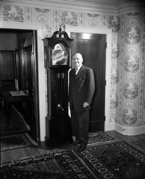 Professor Raymond Dvorak stands beside a grandfather clock in the hallway of his home at 2001 Jefferson Street, decorated with oriental rugs. Mr. Dvorak, director of the U.W. bands who successfully resumed his job after losing his right arm and suffering leg injuries in a train accident, was named head of the Handicapped Day committee. The clock was given to him by the LaCrosse Civic band. He had often guest-conducted the band whose director, Frank Italiano, was an alumnus of the university.