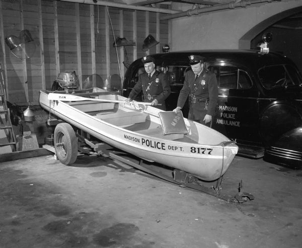 Two uniformed police officers are inspecting a new 14-foot boat with a 15-horsepower outboard motor resting on a boat trailer inside the police garage. In the background is a Madison police ambulance. The new boat was the smallest and most transportable of the police force's three boats.