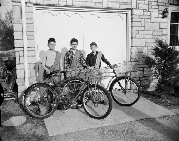 Three paper carriers for the Wisconsin State Journal wear their delivery bags and stand with their bikes against a garage door. The boys are brothers, the sons of Clarence and Catherine Kuehn. They are, from left: Donald, Robert, and Thomas.