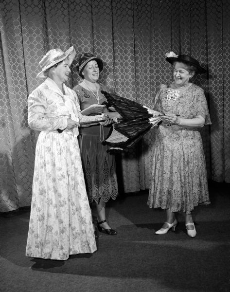 One of a series of scenes from the dramatic production "Down Memory Lane," presented in celebration of the 60th anniversary of the founding of the Madison Woman's Club. Irene Peterson wears a 1908 period dress and shows her black shirred parasol to the other club members. Caroline Homan (center) wears a dress from the 1920's and Edith Johnson wears a costume from 1932.