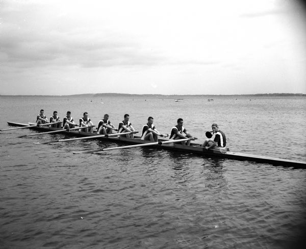 Eight rowers and a coxswain of the University of Wisconsin varsity crew sitting with their oars resting on the water's surface awaiting a start command.