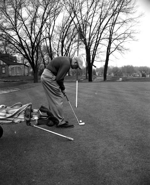George Vitense, a golf professional at Nakoma Golf Club, demonstrates one of seven selected golf etiquette "don'ts" as he misuses his pull cart on the putting green. Part of the clubhouse can be seen in the background on the left.