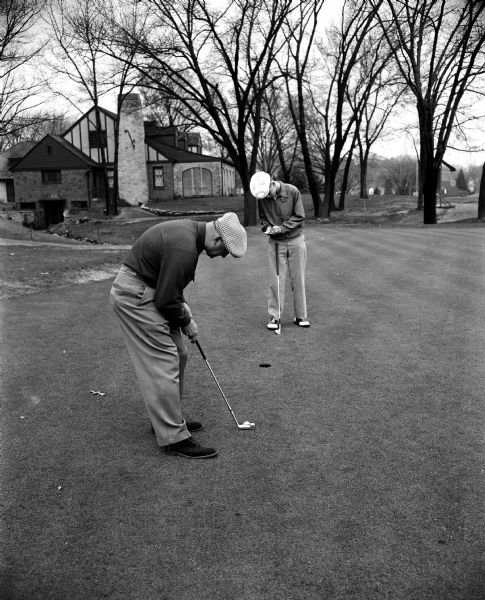 George Vitense, a golf professional at Nakoma Golf Club, lining up a putt while another unnamed golfer is demonstrating "bad" golf etiquette. The Nakoma clubhouse is in the background.