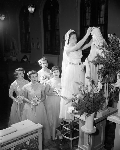 Jacqueline Porter is wearing a long veil and is standing atop a step stool while placing a flower crown on the head of a St. Mary statue inside an unnamed Madison Catholic church. She is attended by, left to right: Helene Baggot, Marilyn DeMuth, Virginia Schmitt, and Mary Verhagen.
