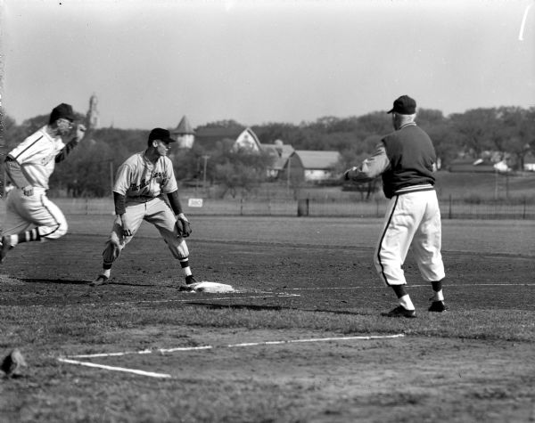 University of Wisconsin Badger Mel Gaestel rounds first base and scores Wisconsin's second run as the Northwestern Wildcat first base man awaits a throw. The view includes a base umpire (right) and the University of Wisconsin barns and a wooded hill in the background.