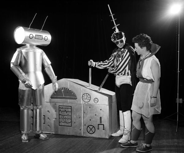 Actors present a University of Wisconsin Medical School junior class skit during Field Day for the medical students. In this skit, "Medirma of the Future," Walter Schwartz, (left) is a robot orderly; Lionel Thatcher is the doctor, and Margaret Thatcher is the student. Their attention is focused on a television X-ray diagnostic machine. 
Field Day is an annual event in which research papers are presented, and various departments have exhibits on their research work, culminating in a banquet and skits by members of the junior class.