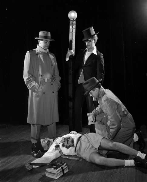 Actors present a University of Wisconsin Medical School junior class skit during Field Day for the medical students. This skit "Willie and the Dragonette," is a play on the TV program "Dragnet." Portraying the characters are, from left: Kenneth Sachtjen, Eugene Yurich, and Lawrence Lund. Ralph Whaley is the "victim" lying on the sidewalk. 
 
Field Day is an annual event in which research papers are presented, and various departments have exhibits on their research work, culminating in a banquet and skits by members of the junior class.