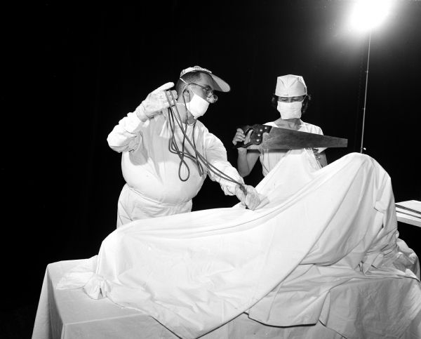 Actors participate in a University of Wisconsin Medical School junior class skit during Field Day for the medical students. This skit, "Mr. Magoo is Called to Surgery," satirizes an operation and surgical techniques. Charles Fitch portrays Mr. Magoo, preparing to operate on a patient. The assisting nurse is portrayed by Elizabeth Smithwick.
 
Field Day is an annual event in which research papers are presented, and various departments have exhibits on their research work, culminating in a banquet and skits by members of the junior class.