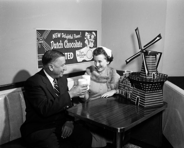 Alice in Dairyland and an unidentified man toast glasses of chocolate milk while seated in a luncheon booth at Rennobahm's drug store. An advertising poster for Dutch chocolate milk with Holland themed symbols hangs on the wall behind them and a model of a Dutch windmill decorates their table.