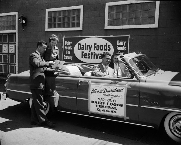 Alice in Dairyland, grand marshall of Madison's Dairy Foods Festival, sits in the back of a convertible parked in front of a building.