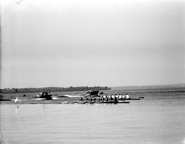 Two nine-men rowing crews racing on Lake Mendota as motor boats full of spectators are looking on. It was the Harvard crew's first visit to Madison and they won the one and 3/4 mile race by 1.3 seconds. University of Wisconsin's Picnic Point is in the background.