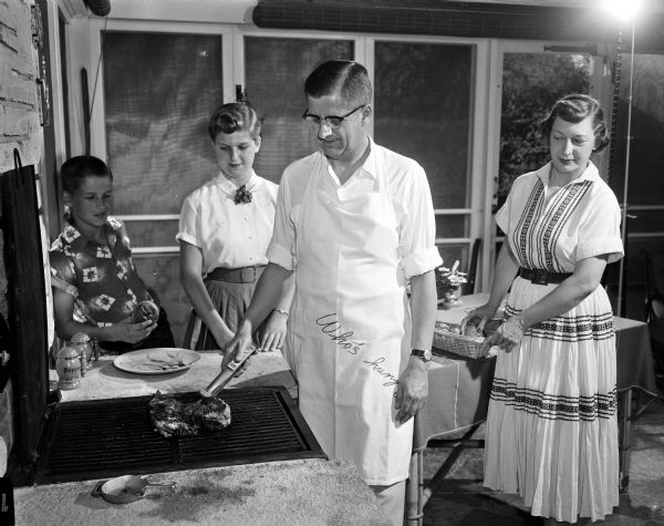 Stanley V. Kubly preparing a steak on a grill while his family is looking on. Watching, from left to right, are Dan, 12; Trudi, 17; and Stan's wife, Evelyn. Stanley is wearing an apron with stitching that reads: "Who's hung[ry?]."