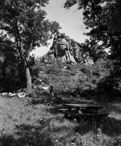 Two people walk near a large rock formation in a wooded area with a picnic table in the foreground. The occasion was the opening of a new wayside park located along Highway 92 between Mt. Horeb and Mt. Vernon. It was named Donald Rock Wayside Park in honor of John S. Donald. The plaque reads: "The site was bequeathed by Mr. Donald, who died in 1934. He served the state as senator and secretary of state, and was a sponsor of the State Highway Aid law of 1911, which formed the framework for the state's highway system."