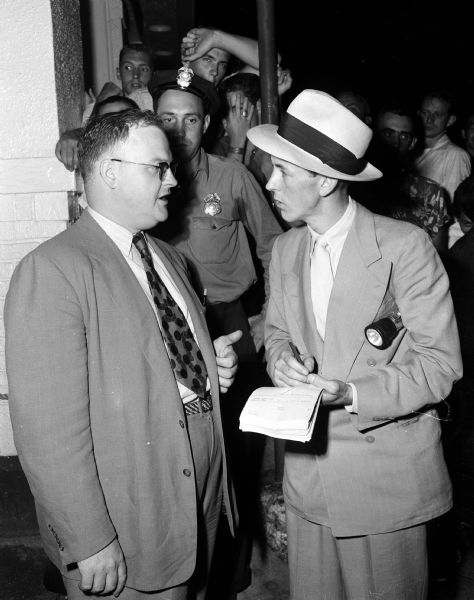 James Quinn, witness of the events leading to the murder and suicide of Doris Russ and Guy Russ, talks to either police investigator John Henry or newspaper reporter James Robinson. The investigator (right) holds a pencil and note book in hand and a flashlight under his arm. Behind him a uniformed police officer is standing in front of a small crowd of onlookers.