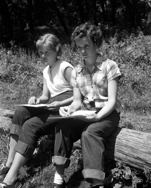Two campers at Camp Maria Olbrich, a YWCA camp on the shore of Lake Mendota near Fox Bluff, sit on a log while making sketches. The girls are Susan Meyer (left), daughter of Mr. and Mrs. Arthur J. Meyer of 1302 Nishishin Trail, and Gretchen Huebner, daughter of Mr. and Mrs. Delmar Huebner, 519 South Orchard Street.