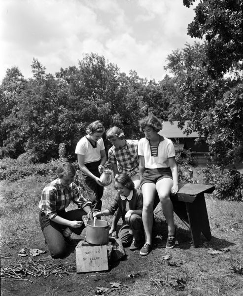 Five girls prepare a meal around a campfire at Camp Maria Olbrich, a YWCA Teen Camp on the shore of Lake Mendota near Fox Bluff.
Left to right are: Carol Tygum, daughter of Mr. and Mrs. Eldred Tygum, 4105 Hillcrest Drive, one of the counselors; Pamela Reis, daughter of Dr. and Mrs. Louis N. Reis, 701 Seneca Place; Nancy Swanson, daughter of Mr. and Mrs. Charles L. Swanson, 4901 Tonyawatha Trail; Lana Lokken, kneeling, daughter of Mr. and Mrs. Lloyd E. Beckett, 3 Frances Court; and Faye Kaker, daughter of Mr. and Mrs. Paul C. Fakler, 1347 Jenifer Street.