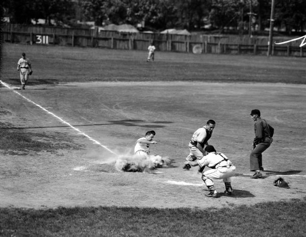 Right fielder Bob Kessenich slides into home plate in a cloud of dust with Waunakee's sixth run in its 7-0 victory over Sauk-Prairie at Sauk City. Catcher Karl Wenzel (capless) of Sauk-Prairie and catcher John Taylor of Waunakee are in the foreground.