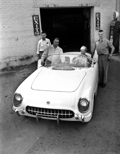 Larry Jacobson, the 1953 Madison Soap Box Derby champion, sitting in the Chevrolet Corvette which will carry him at the head of the 1954 parade. He is wearing the star-studded helmet and shirt he won in Akron, Ohio. Members of the Hult's garage crew, a race sponsor, are standing around the car. They are, left to right, Willis Powers, George Lambrect, and Lowell Hammer. They are at 627 East Mifflin Street and, in the background, is a service garage exit, a gas pump, and a poster.