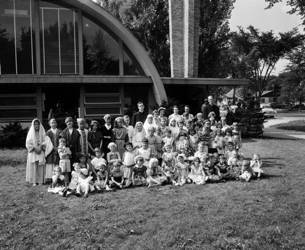 Portrait of over 50 students and faculty posing on their last day of bible school in front of their church at 4011 Major Avenue. The church is a mid-century building with half-barrel shaped roof and walls. Some of the children are wearing costumes for a play entitled "The Raising of Lazarus."