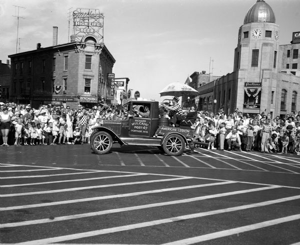 An old truck operated by the Richard Mulcahy Post 47 of Portage spraying water on the crowd at the American Legion parade around Capitol Square. The view is looking toward King Street on the Capitol Square.