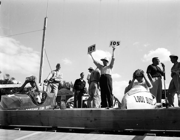 View of the starting line scene at the 1954 Madison soap box derby. Racers Ted Scherbert of Portage (left) and Gerald Flentje of Lodi adjust their helmets while Tom Riggs of radio station WKOW holds up cards. At the right is veteran master of ceremonies Dr. William Rundell holding a microphone.