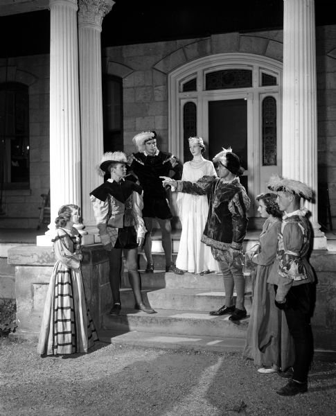 Costumed actors and actresses from the Mineral Point Players stand on the steps of the Gundry Mansion while enacting a scene from Shakespeare's "A Midsummer Night's Dream". Left to right: Marlyn Adams, James McNeil, Milton Nelson, Pat McGettigan, Gerald Adams, Gertrude Dalles, and Jerry Whitford.