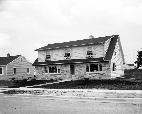 View across street towards the Ernie Terhall house in at 560 Kelly Street. It is a two-story house fronted with Lannon stone and siding. There are two picture windows on the first floor and a full-length three window dormer on the second floor.