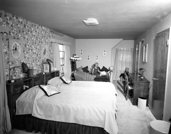 Portrait of Sharon Terhall in her bedroom, complete with two twin beds with reading lamps on headboards, a desk, a wallpapered wall, and upholstered chairs.