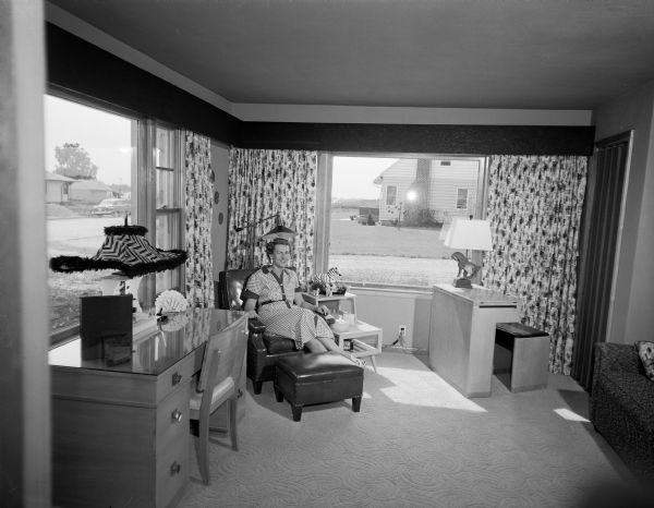 Mrs. Terhall is relaxing in an easy chair in the den of her family's new home. Two picture windows, identical to the two on the front of the house, nearly meet in the corner behind Mrs. Terhall. Neighboring houses are visible through the two windows. The room also includes draperies, a desk, and other furniture.