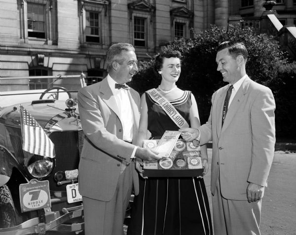 Edward Hansen, left, standing in front of his 1918 Biddle automobile at the Wisconsin State Capitol prior to leaving for Scotland to participate in the Anglo-American Vintage Car Rally. Alice in Dairyland, Mary Ellen McCabe, presents him with a package of Wisconsin cheese and Arthur Wichern, public relations director for the American Automobile Association, gives him a map of Scotland.