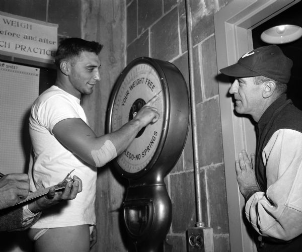 University of Wisconsin football fullback Alan Ameche stands on a scale and points to his 210-pound weight as coach Ivan Williamson looks on.