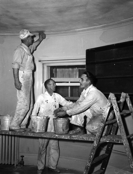 Three construction workers are dressed in white work clothes while working with sponges, pails, and a scaffold, and standing by an open window. They include Ed Woerpel, Sylvester Schmit and Ernest Parker; the men are donating a half day's pay to the United Givers' Fund.