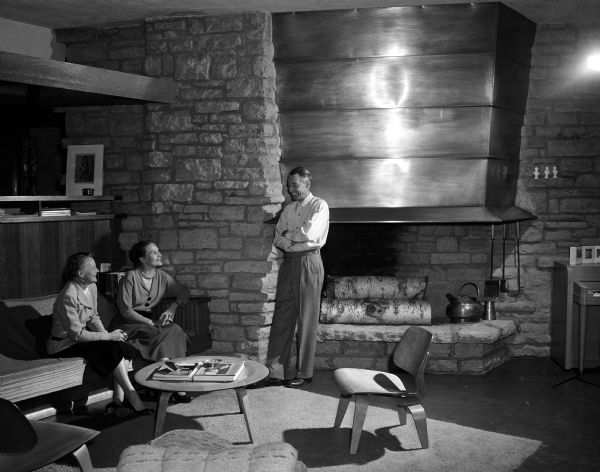 Mr. William Kaeser stands in front of a large fireplace at his home at 3467 Circle Close, one of the houses on the League of Women Voters' House Tour. Mrs. Porter Butts (left) and Bonnie Kienitz are seated on the couch.