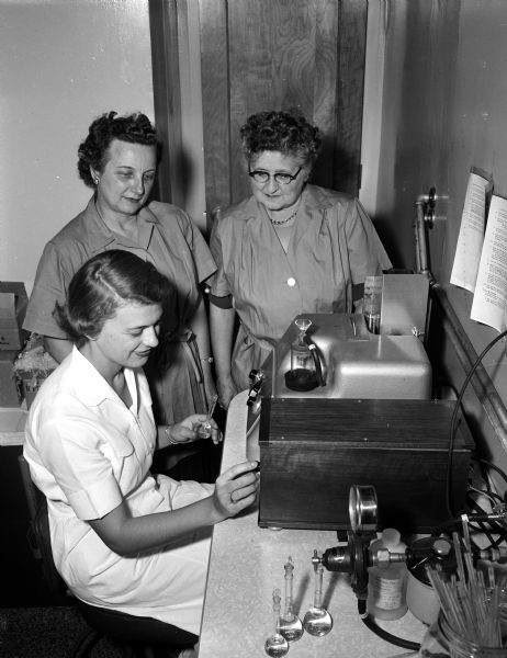Two volunteer workers of the Madison General Hospital auxiliary, Mrs. John F. Mullen (left), West Point, and Mrs. William W. (Melva) Marling of 302 Walnut Street, watch Miss Miriam Anderson, 310 South Orchard Street, operate a flame photometer. Anderson was a medical technician at the hospital and the photometer was a new piece of laboratory equipment.
