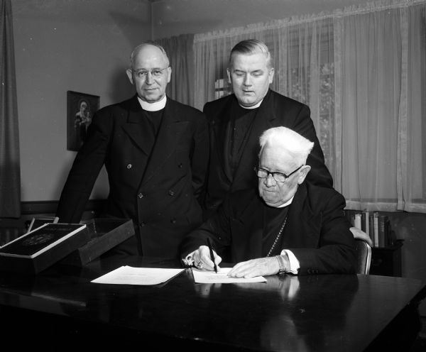 The Most Reverend William P. O'Connor, Catholic bishop of Madison, signs the letter in which he asks Pope Pius XII for formal permission for the Redemptorist Fathers to enter his diocese to erect their new minor seminary near Albion, Wisconsin. Looking on are the Reverend William H. Reintjes, C.SS.R., St. Louis, Missouri, first assistant to the St. Louis provincial of the society, left, and the Reverend Francis O'Neill, C.SS.R., rector of the Redemptorists' major seminary at Oconomowoc, Wisconsin.