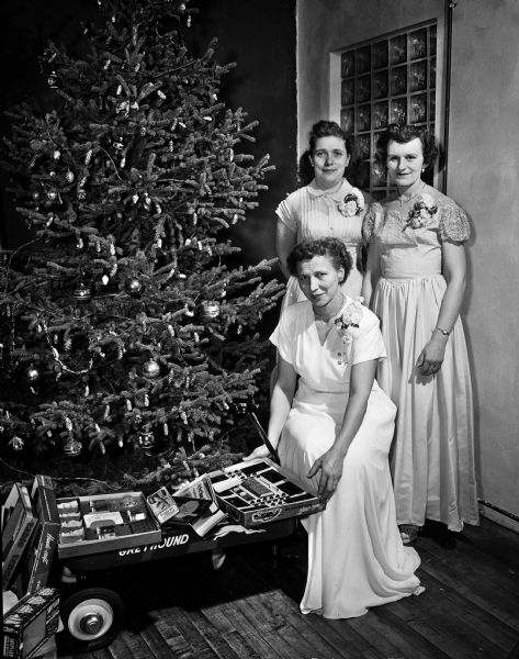 Women of the Moose display Christmas gifts to be given to children at Mooseheart, Illinois and for the aged at Moosehaven, Florida. Darlene Frazier (Mrs. Albert Frazier) stands beside the Christmas tree and, standing left to right, are Mrs. John Steel and Mrs. Charles Pollock.