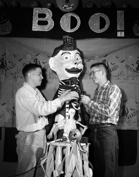 Kent Kildow and Larry Brown are shown adding an extra loop of paper ribbon to the spooky figure on the band stand. Other paper decorations include skeletons, cats, jackolanterns, and a large "BOO!" sign.
