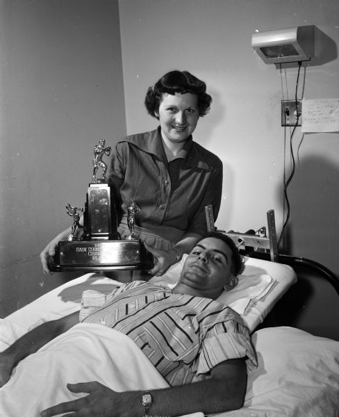 Eddie Paulus, outfielder for the Plain, Wisconsin baseball team, lies in a hospital bed at St. Mary's Hospital in Madison after a collision with a catcher at home base. His wife, Viola, sits next to him with the Sauk County Baseball League championship trophy.