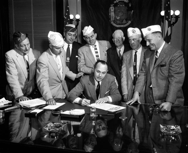 Governor Walter Kohler signs a proclamation making November 11, 1954 the first Veterans Day in Wisconsin. Looking on, left to right, are: Francis Lorbecki of Watertown; Morris Pendleton, A.J. Healy, Conrad Lewis, William Gillette, G. Earl Heath, all of Madison; James Martineau of Oconto.