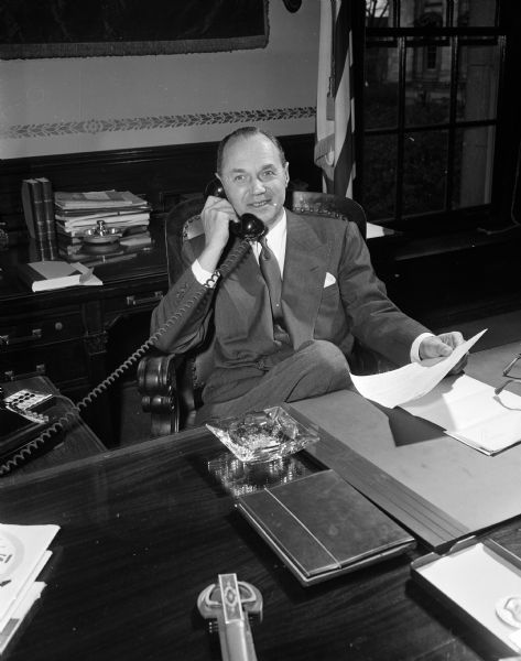 Wisconsin Governor Walter Kohler sits at his desk in the Wisconsin State Capitol executive office while making phone calls to encourage state residents to vote Republican in the upcoming election. He holds a photograph of President Eisenhower.