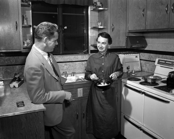 Stewardess Coleen Ogle cooking in the apartment she shares with three other stewardesses. Art Kemp, first officer and co-pilot, is looking on. The photograph was taken to accompany an article about the daily lives of six North Central airline stewardess assigned to Madison as their home base.