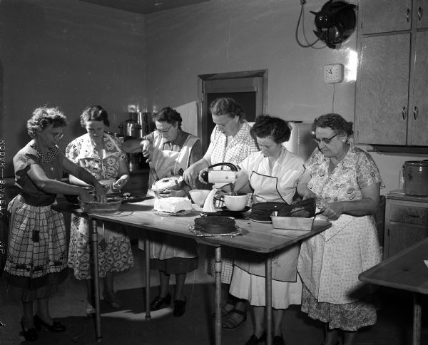 Six women, some wearing aprons, preparing cakes while standing around a high table.  Cakes and a stand mixer are on the table top. They are at Lakeview American Lutheran church at 4001 Mandrake Street off of Northport Drive. Left to right, they include: Amelia Stang, Arvada Kuhlmey, Malinda Koenig, Grace West, Evelyn Lee, and Ernestine Borchers. They are making preparations for their traditional fall lutefisk and chicken dinner.