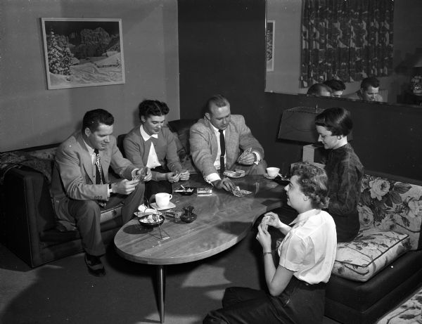 Slightly elevated view of three women and two men playing cards while sitting on Mid-Century couches around a kidney shaped coffee table. On the coffee table are two coffee cups, a cigarette ashtray, and a snack dish.