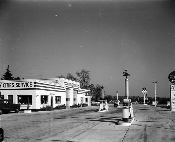Exterior view of a large service station with three groups of gasoline pumps in front, located at 2629 West Broadway in Monona. The building has a sales room, two service bays, and a diner with an "EAT" sign.