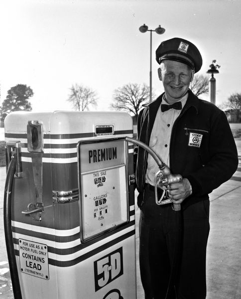 Advertising portrait of Ole Olson. He is wearing a uniform and bow tie with emblem patches on his jacket and cap. He is holding a gas hose nozzle and is standing by a gas pump that is set to 32.9 cents per gallon.