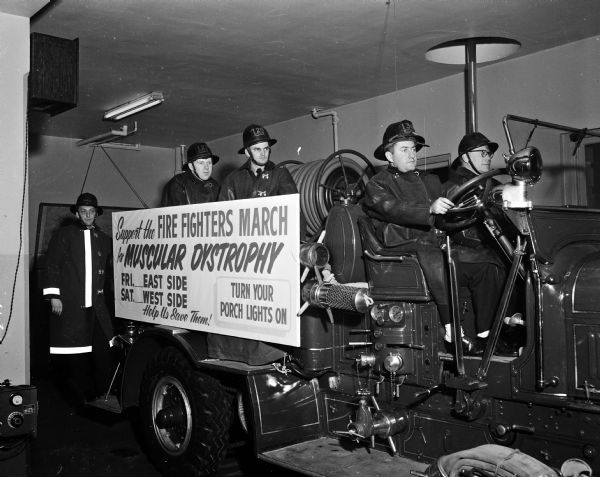 Four firefighters wearing working gear and positioned about a fire truck posing with a sign stating: "Firefighters March on Muscular Dystrophy." On the back of the truck are Jim Shipley and Bill Ferger. Carson Mettel is in the driver's seat and beside him is Walter Ayers. They intended to raise funds over a two night period with a porchlight campaign. In the background is a fireman's pole leading from the second floor to the first floor. They are inside Fire Station #2 at 301 North Broom Street.